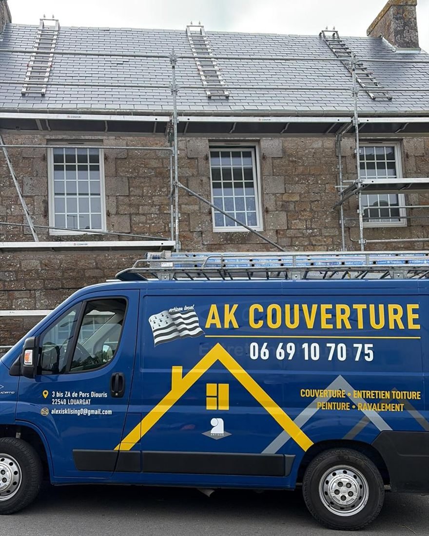 Le fourgon bleu et jaune d’AK Couverture garé devant une maison en pierre dont la toiture en ardoise est en cours de réfection, sous échafaudage